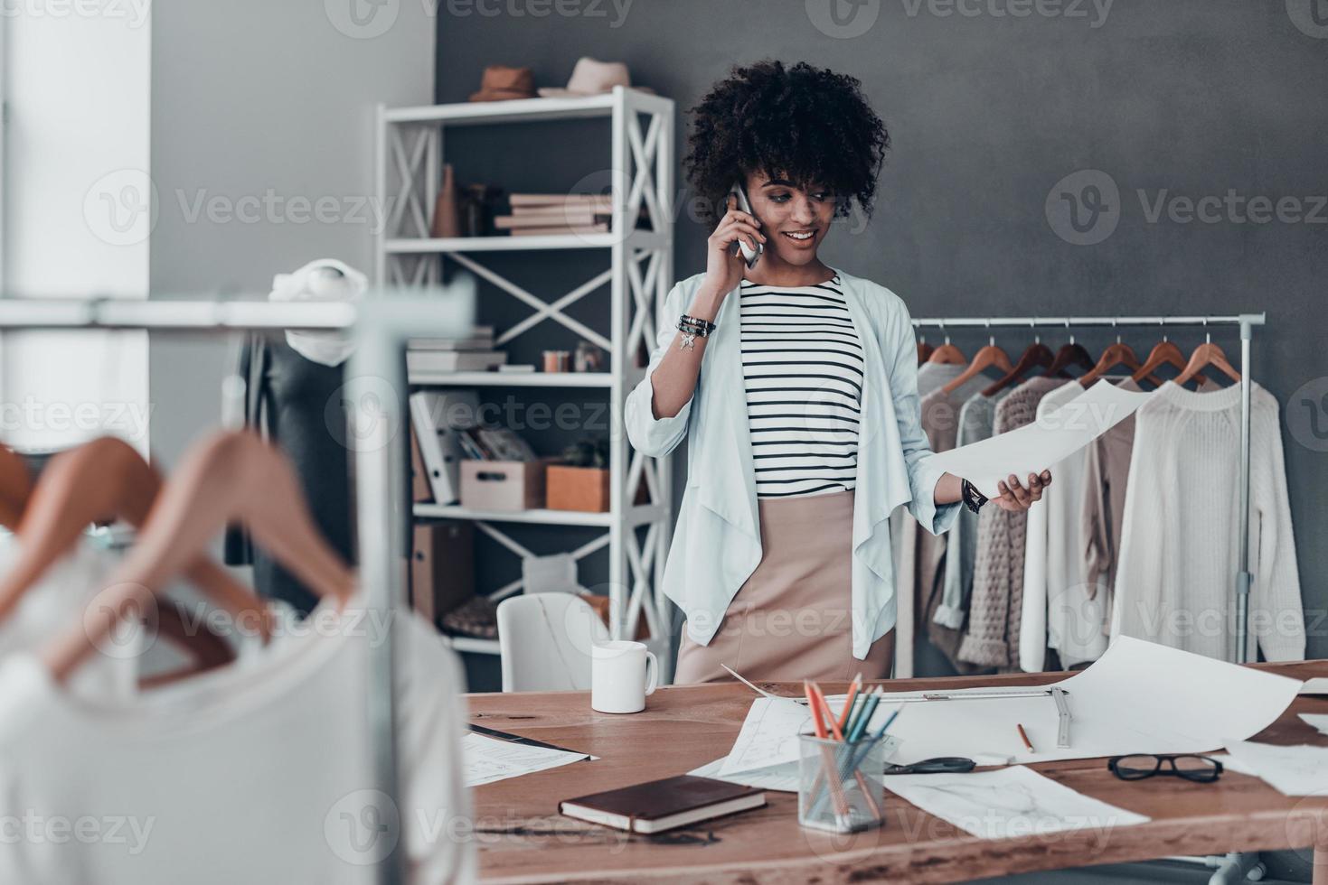 siempre disponible. hermosa joven africana hablando por teléfono inteligente y sonriendo mientras está de pie en su taller foto