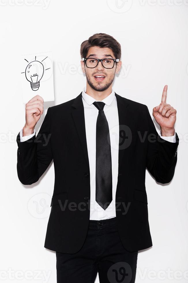 Good idea Excited young man in formalwear pointing up and holding picture of electric lamp while standing against white background photo