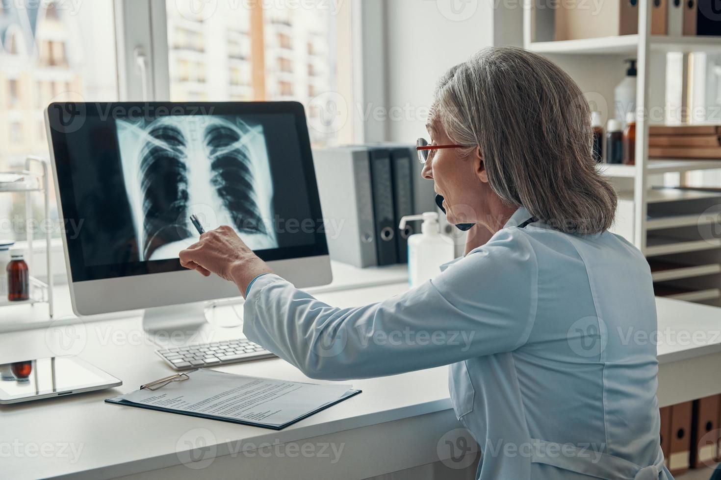 Mature female doctor in white lab coat talking on the phone and examining lungs using computer while sitting in her office photo