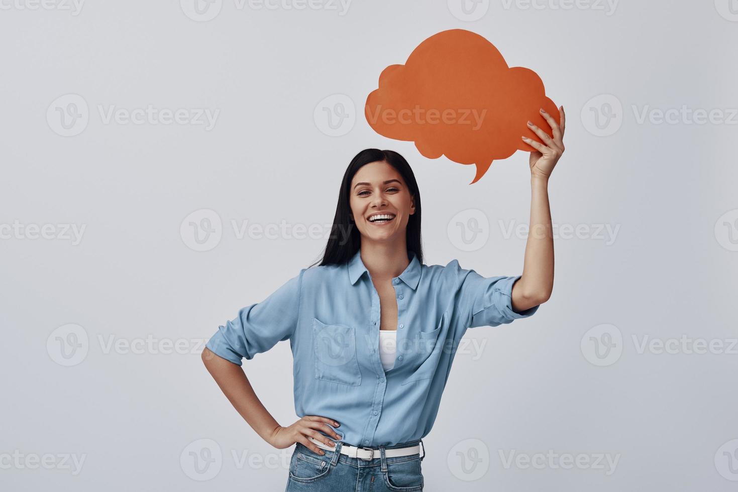 Attractive young woman holding speech bubble and smiling while standing against grey background photo