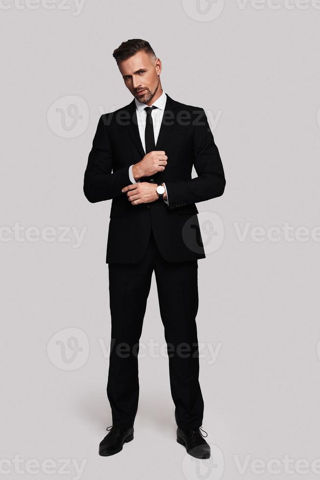 Paying attention to every detail. Full length of handsome young man in full suit adjusting sleeve and looking at camera while standing against grey background photo