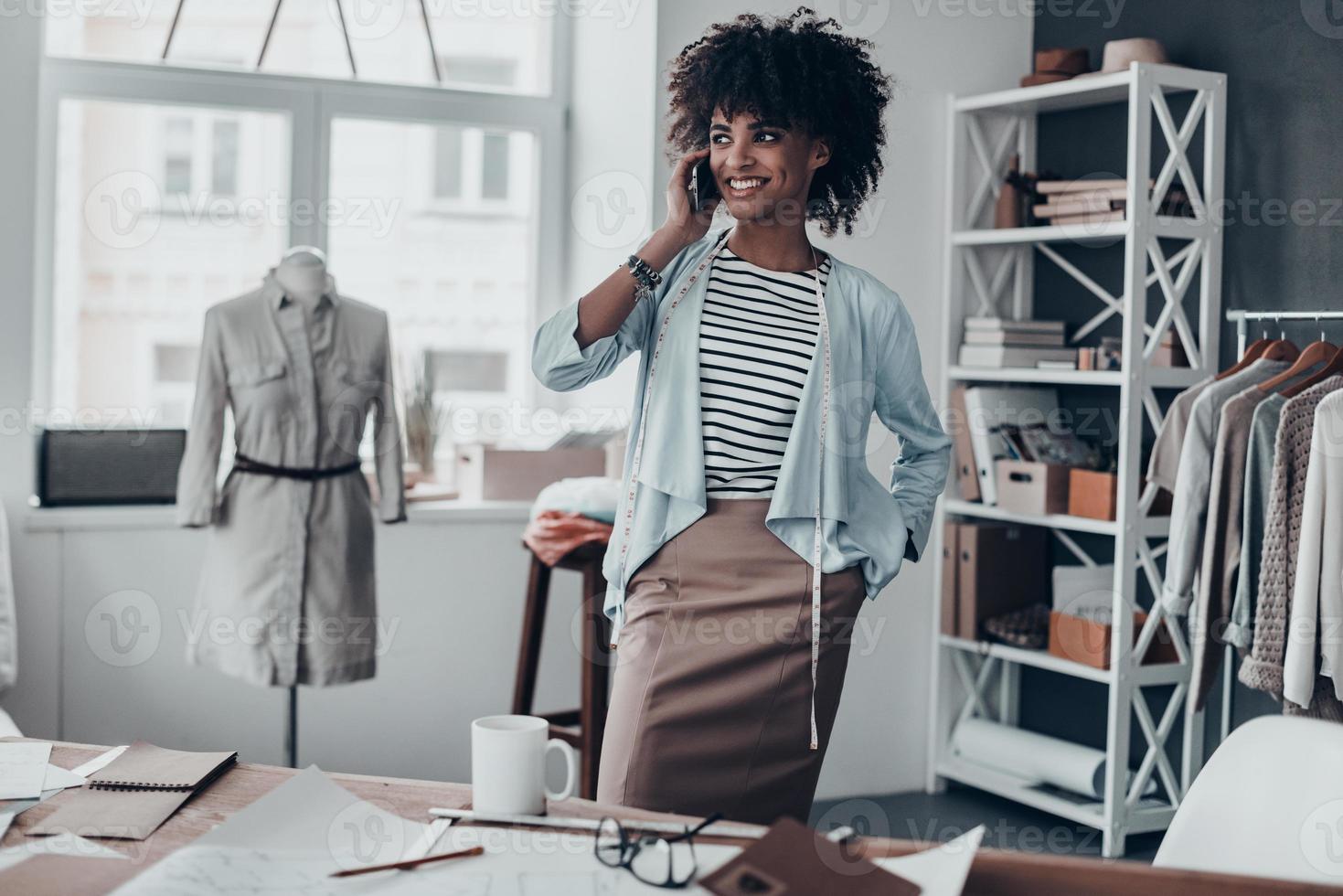 buena charla con el cliente. atractiva joven africana hablando por teléfono inteligente y mirando hacia otro lado con una sonrisa mientras estaba en su taller foto