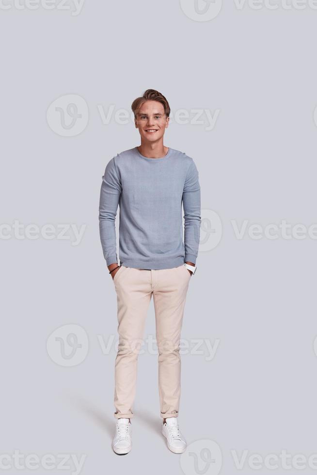 Simply handsome. Full length of charming young man looking at camera and keeping hands in pockets while standing against grey background photo