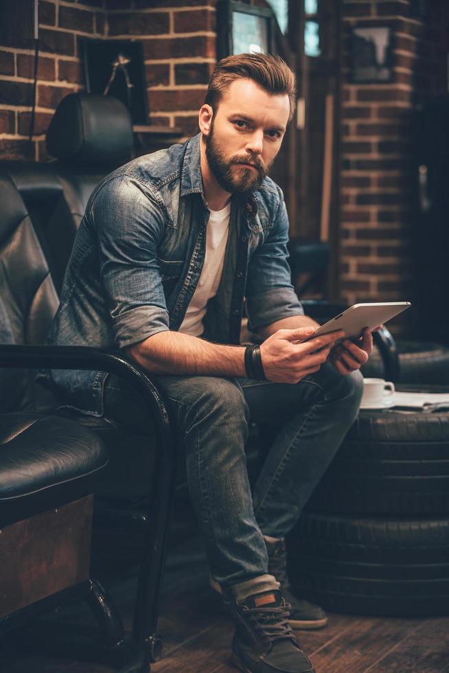 ponerse al día con algunos peinados en línea. apuesto joven barbudo sosteniendo una tableta digital y mirando a la cámara mientras se sienta en una silla cómoda en la barbería foto