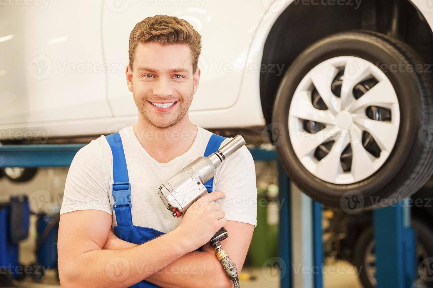 Ready to work. Confident young man holding a pneumatic wrench and smiling while standing in workshop with car in the background photo