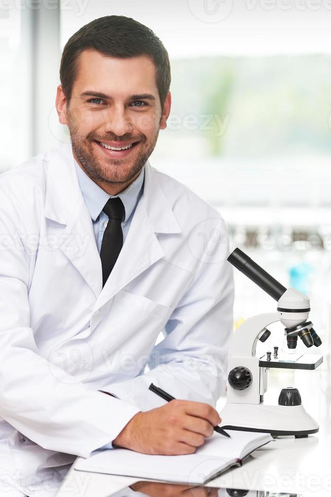 la ciencia es mi vida. joven científico sonriente con uniforme blanco escribiendo en su bloc de notas y mirando la cámara mientras se sienta en su lugar de trabajo foto
