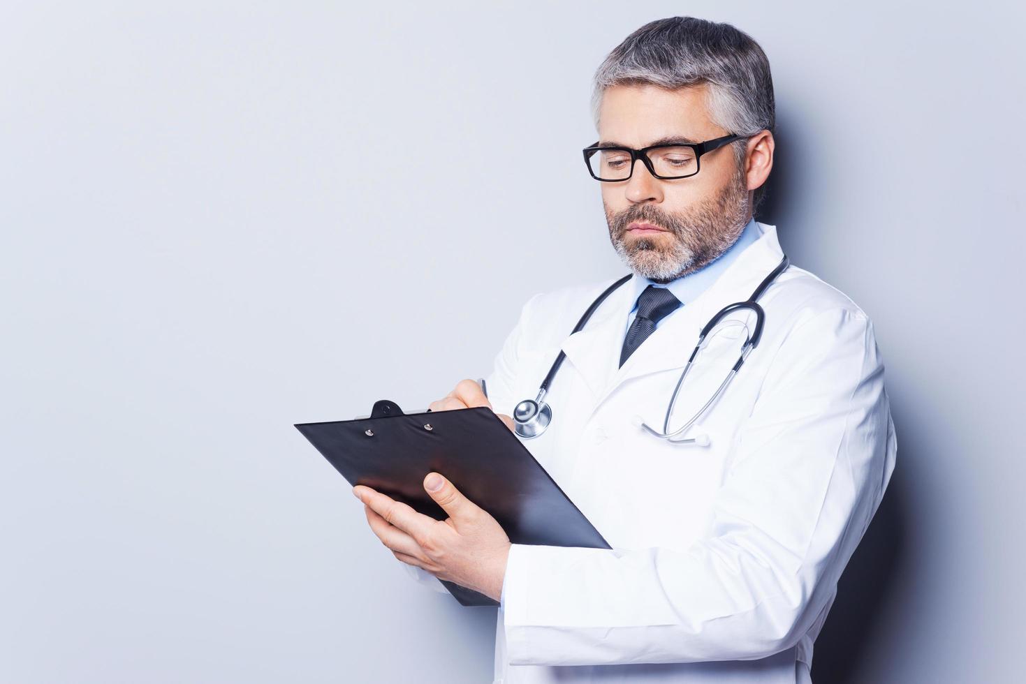 Doctor writing RX prescription. Confident mature doctor writing something at his clipboard while standing against grey background photo