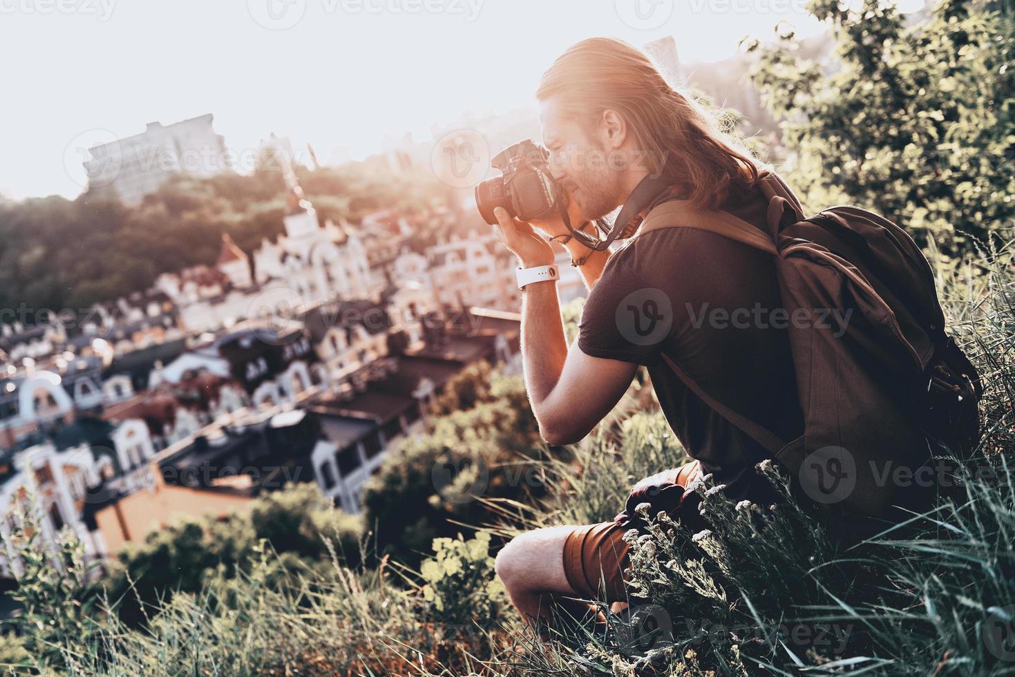 Capturing beautiful view. Young man in casual clothing photographing the view while sitting on the hill outdoors photo