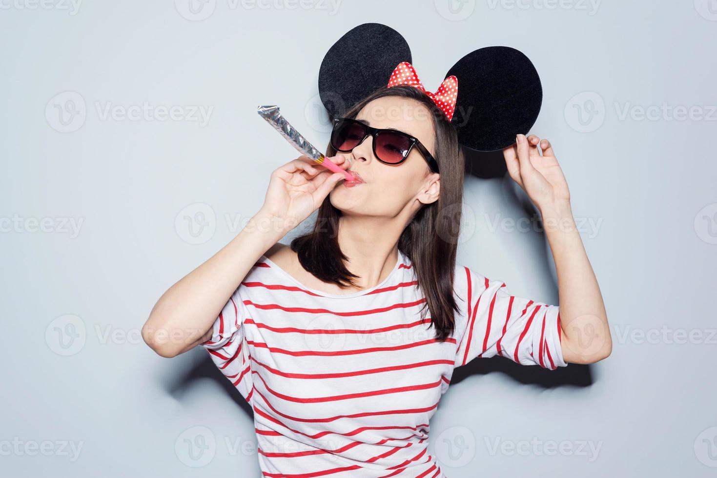 She clearly loves holidays  Playful young woman in glasses and mouse ears blowing in noisemaker while standing against grey background photo