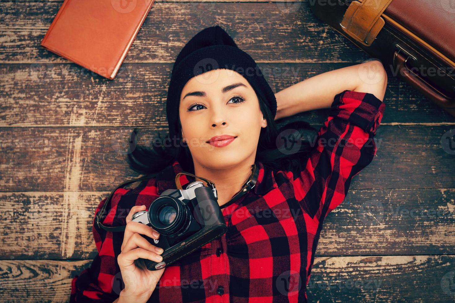 hermosa soñadora. vista superior de una hermosa joven con ropa de cabeza tirada en el suelo y sosteniendo la cámara mientras la maleta y el bloc de notas yacen cerca de ella foto