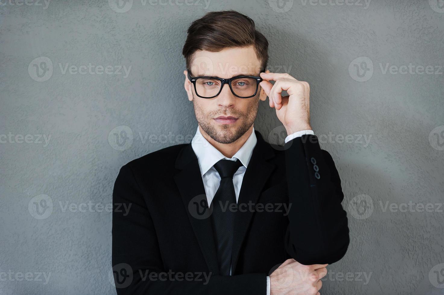 Stylish handsome. Confident young businessman adjusting his eyewear and looking at camera while standing against grey background photo