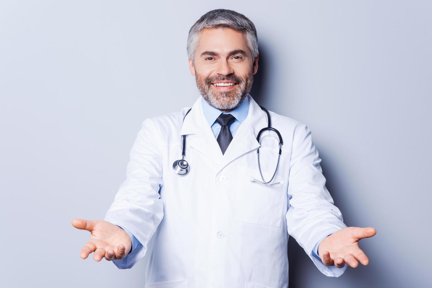 médico alegre. alegre doctor de pelo gris maduro mirando a la cámara mientras estira las manos y sonríe mientras está de pie contra el fondo gris foto