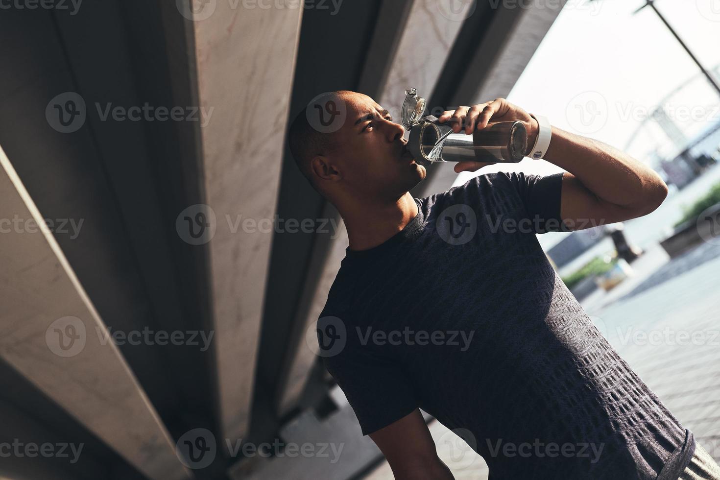Hydrating. Young African man in sports clothing drinking water while standing outside photo