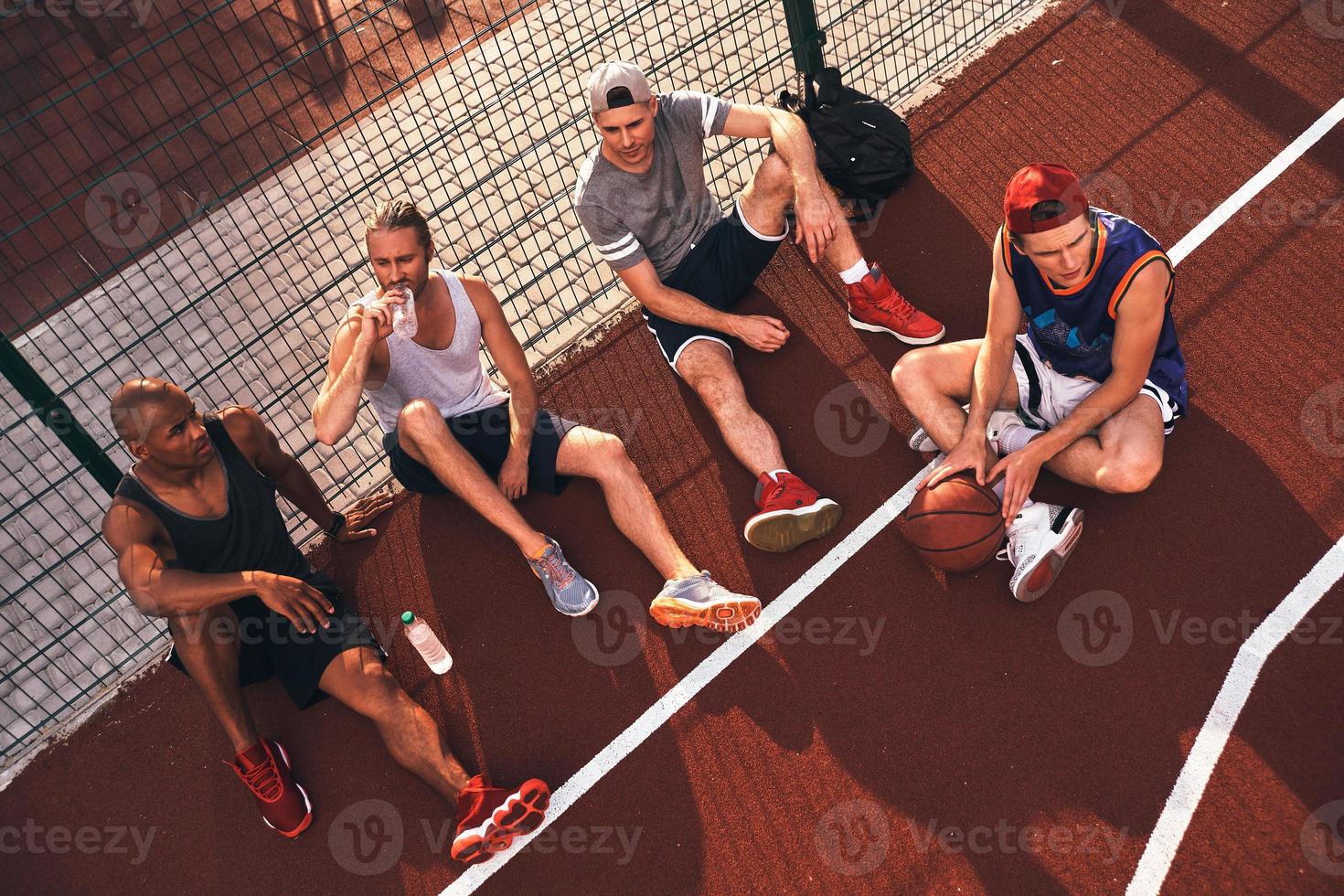 Tired after game. Top view of young men in sports clothing looking away while sitting on the basketball field outdoors photo