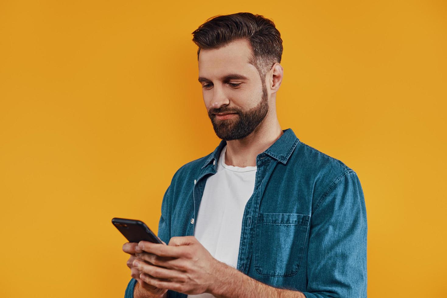 Handsome young man in casual clothing using smart phone and smiling while standing against yellow background photo