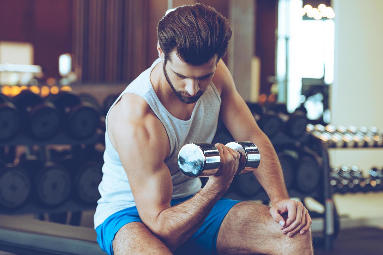 siente la quemadura en el bíceps. hombre guapo joven confiado en ropa deportiva haciendo ejercicio con pesas en el gimnasio foto