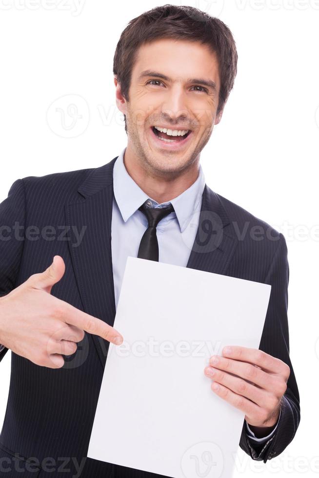 mira a este joven feliz con ropa formal sosteniendo un papel en blanco y señalándolo con una sonrisa mientras está aislado en el fondo blanco foto