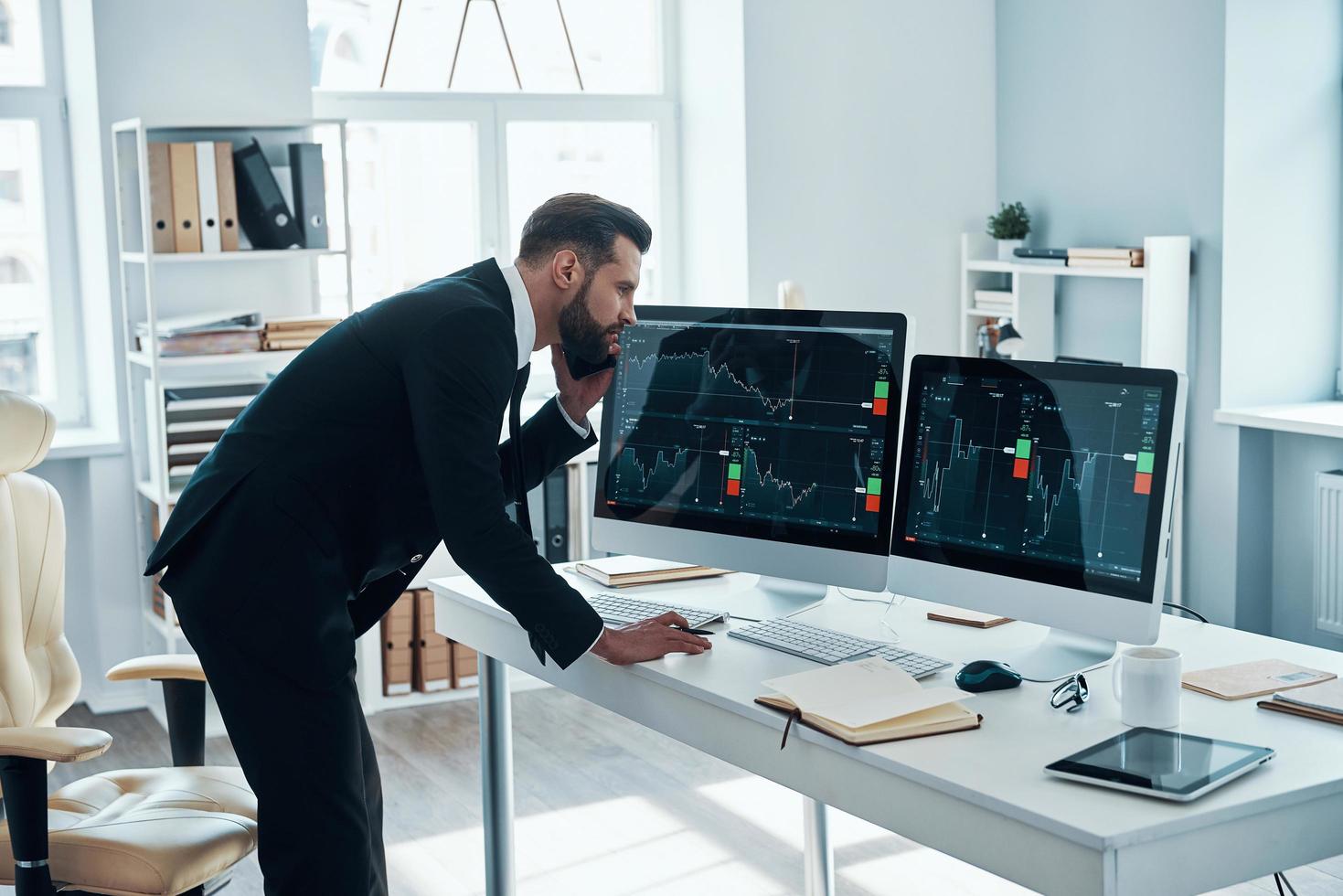 Concentrated young man in shirt and tie writing something down and talking on the phone while working in the office photo