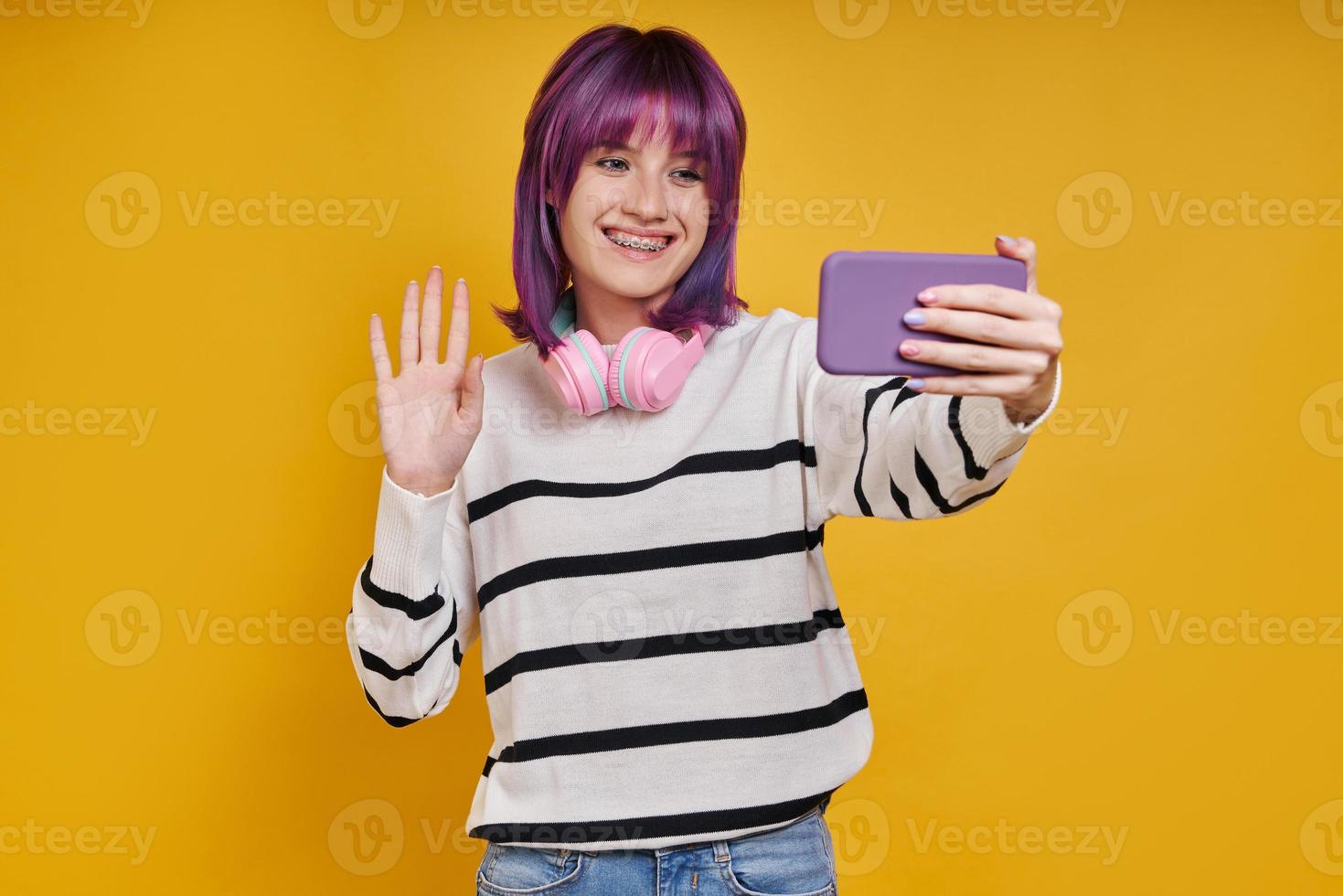 Cheerful young woman having video call while standing against yellow background photo