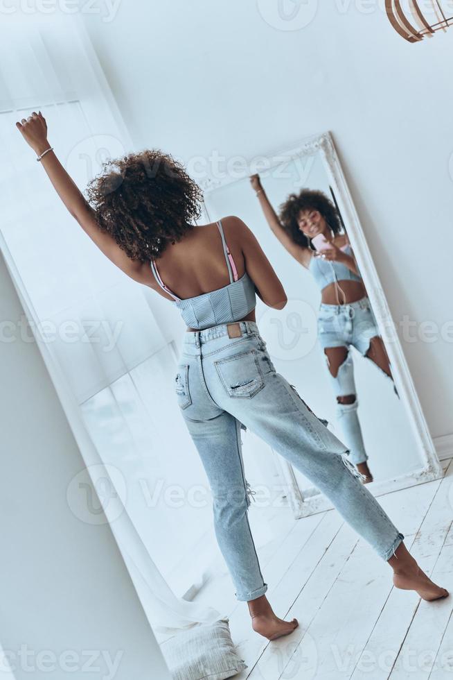 She is a superstar. Full length rear view of beautiful young African woman listening music and smiling while taking selfie at home photo