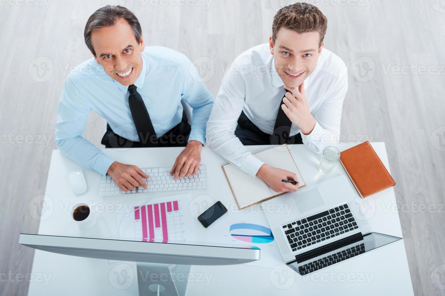 lo de siempre. vista superior de dos alegres empresarios con ropa formal trabajando y mirando la cámara mientras se sientan en el lugar de trabajo foto