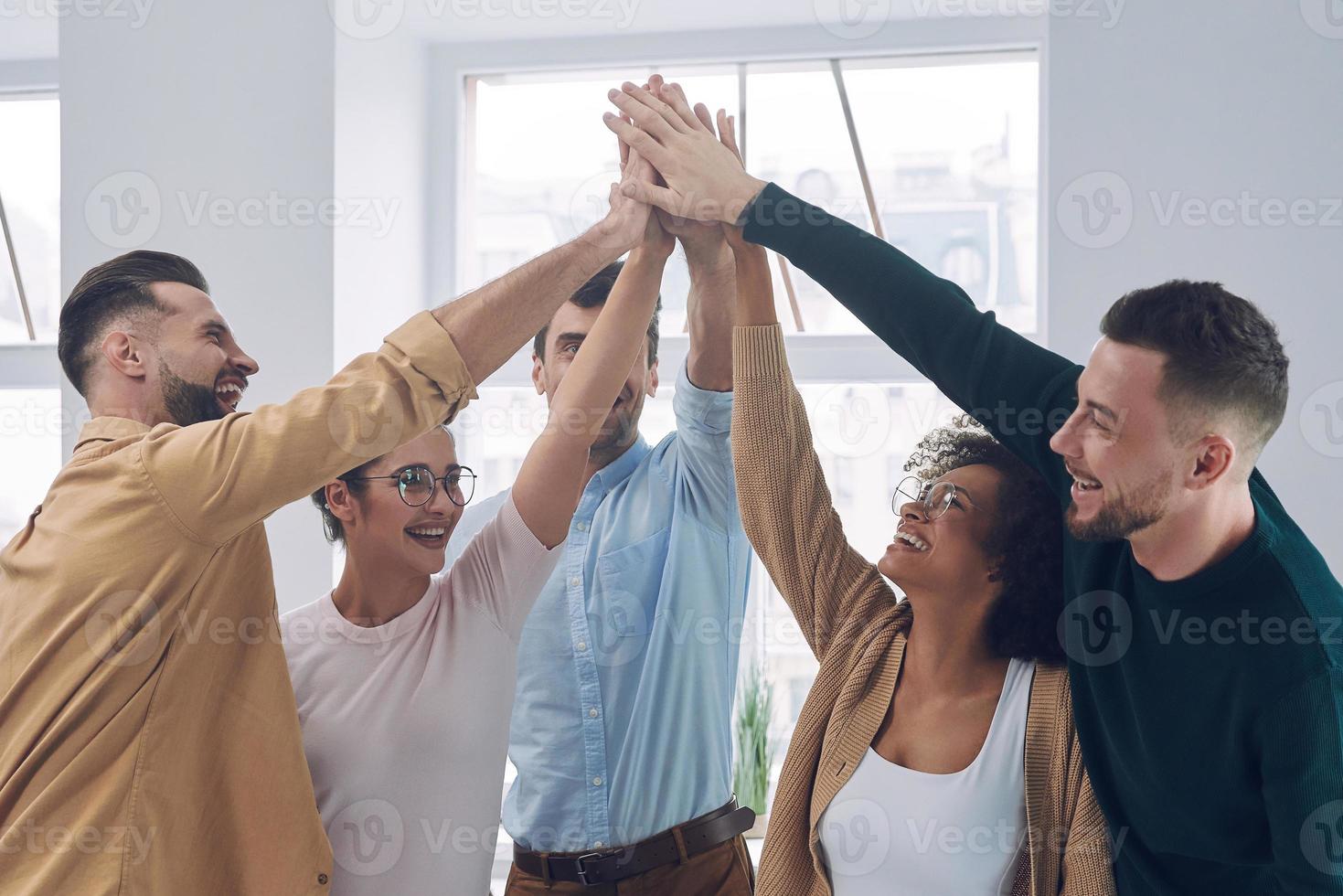 grupo de jóvenes felices con ropa informal inteligente tomados de la mano y sonriendo mientras se reúnen en la oficina foto