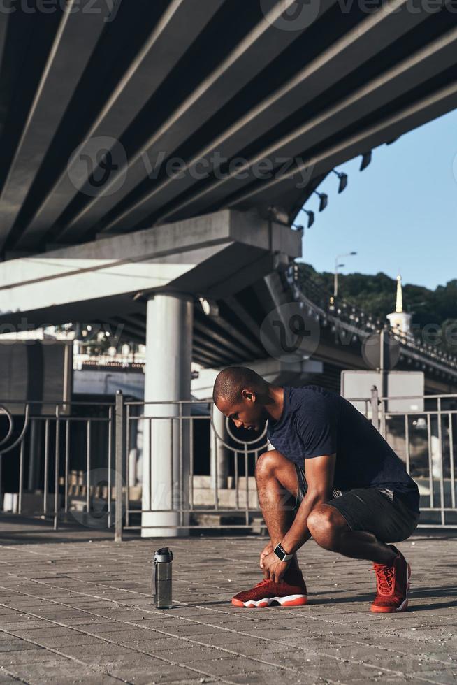 Getting ready. Handsome young African man in sports clothing tying shoes while standing outdoors photo