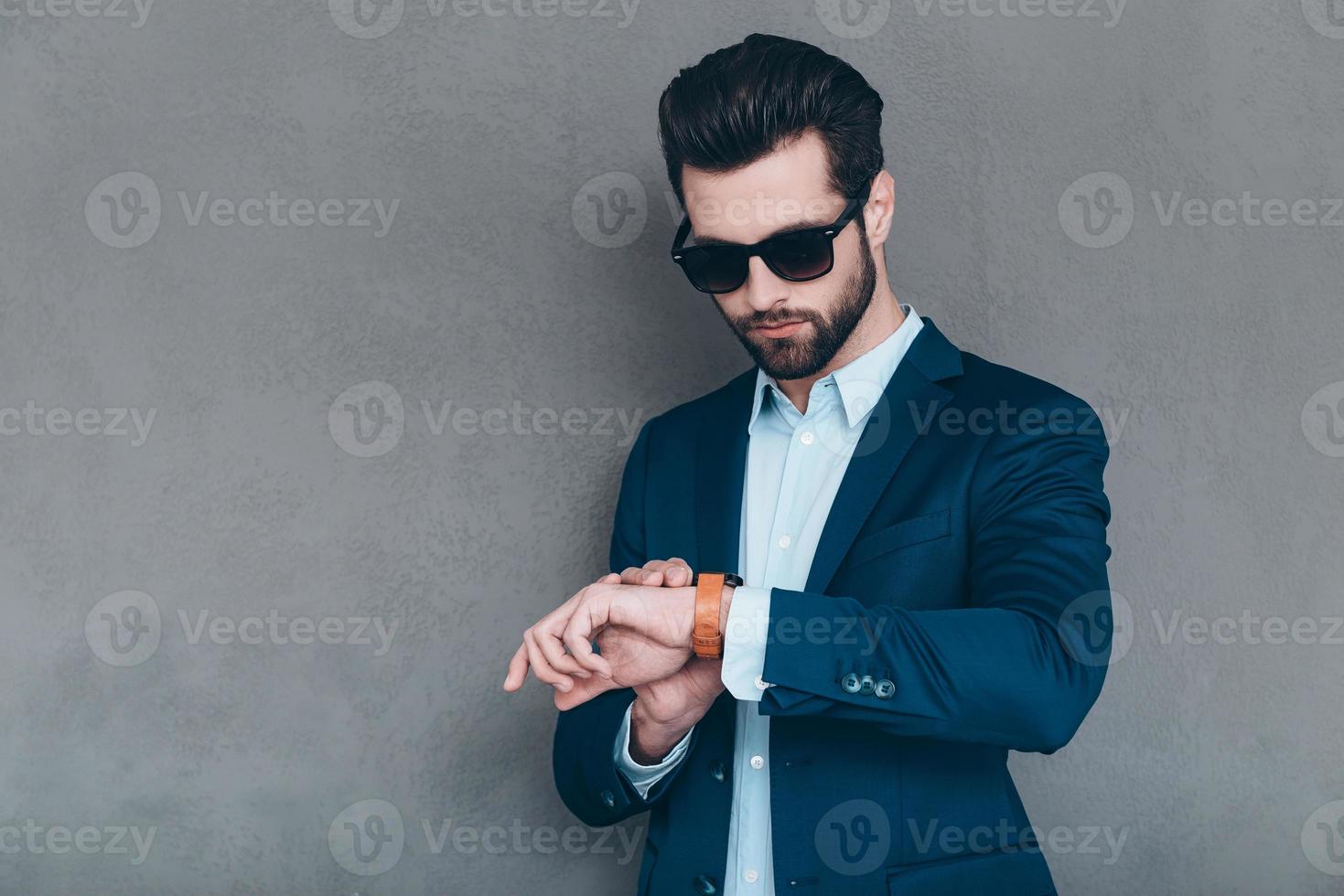 Having a date in one hour. Handsome young man wearing sunglasses and looking at his wristwatch while standing against grey background photo