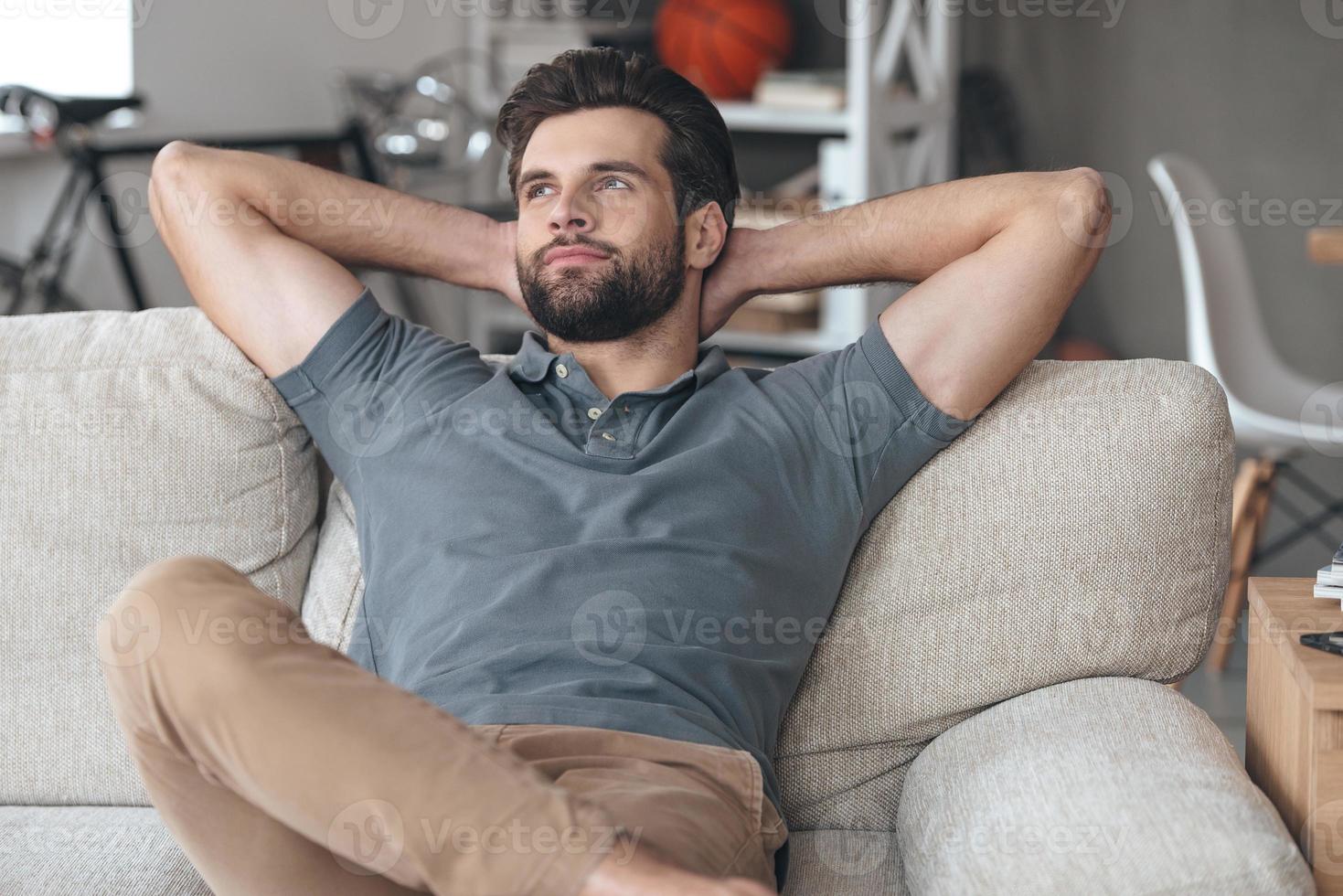 Time to gather all ideas in his head. Handsome young man keeping hands behind his head and looking thoughtful while sitting on the couch at home photo