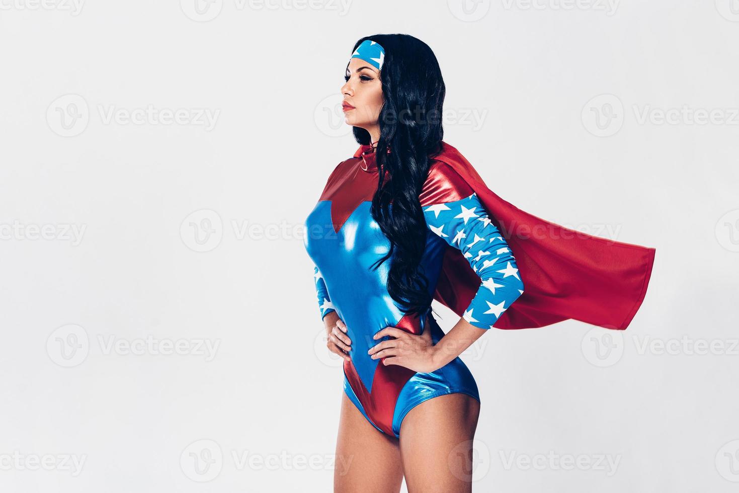 Ready to save the world. Side view of beautiful young woman in superhero costume keeping hands on hips while standing against white background photo