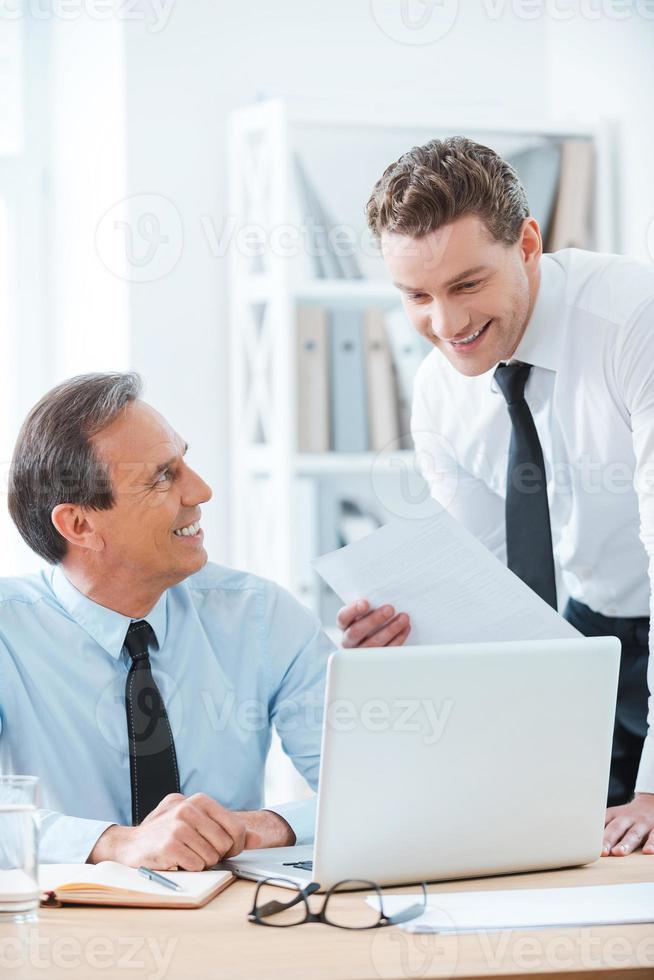 Sharing business ideas. Smiling mature businessman sitting at his working place while young man standing near with documents photo