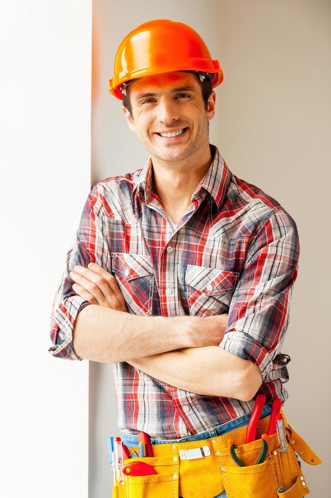 reparador confiado. apuesto joven manitas con casco apoyado en la pared y sonriendo a la cámara foto