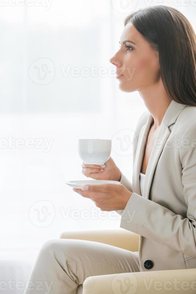 Coffee break. Side view of thoughtful young businesswoman in suit drinking coffee and looking away while sitting at the chair photo
