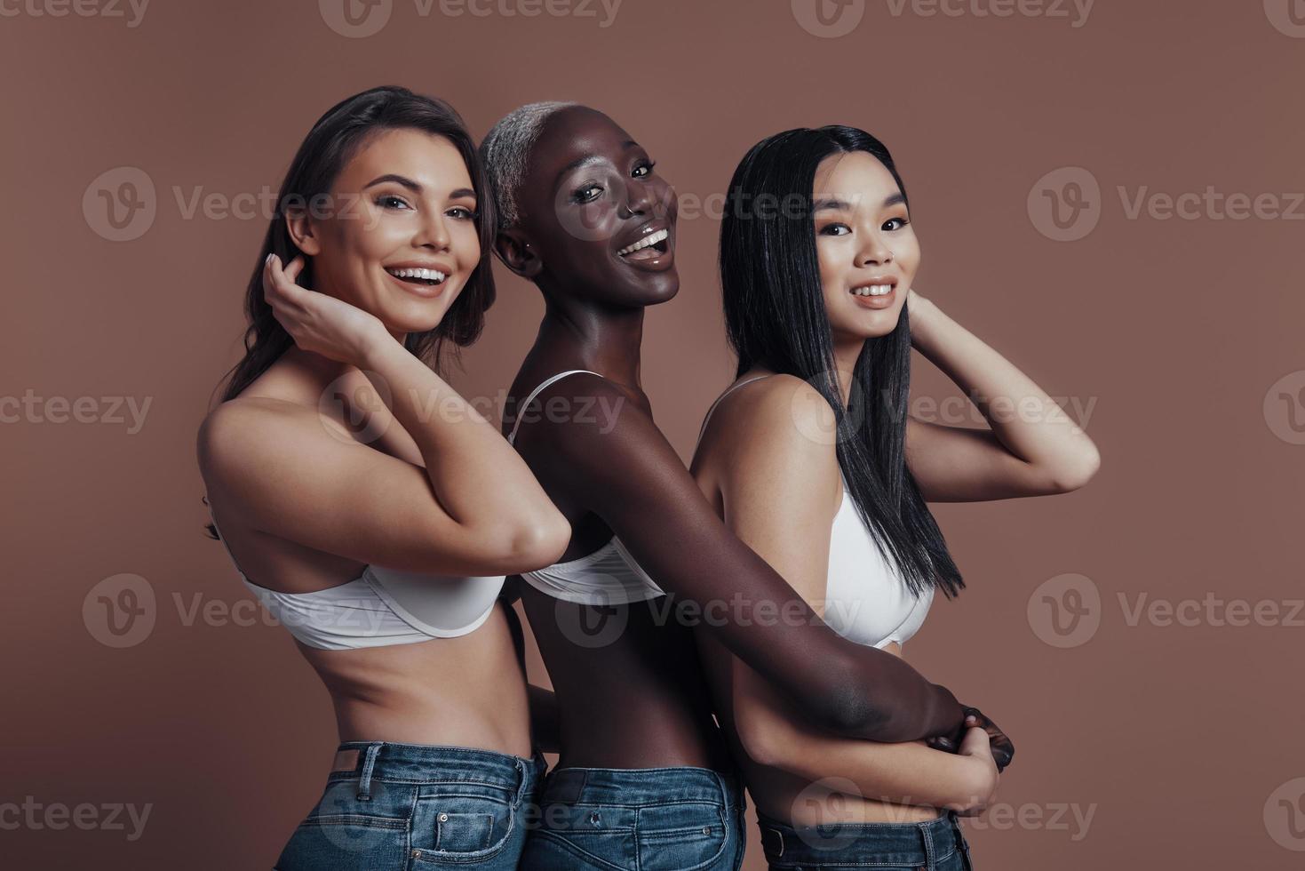 Cheerful beauties. Three attractive young women looking at camera and smiling while standing against brown background photo