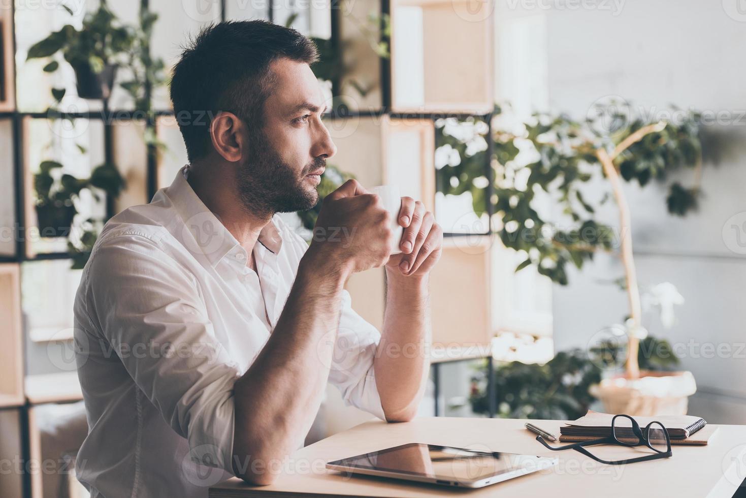 Waiting for inspiration. Side view of thoughtful young man holding cup of coffee and looking away while sitting in cafe photo