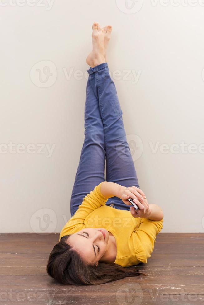 belleza con teléfono móvil. bella joven sosteniendo un teléfono móvil mientras está tirada en el suelo y apoyando las piernas en la pared foto