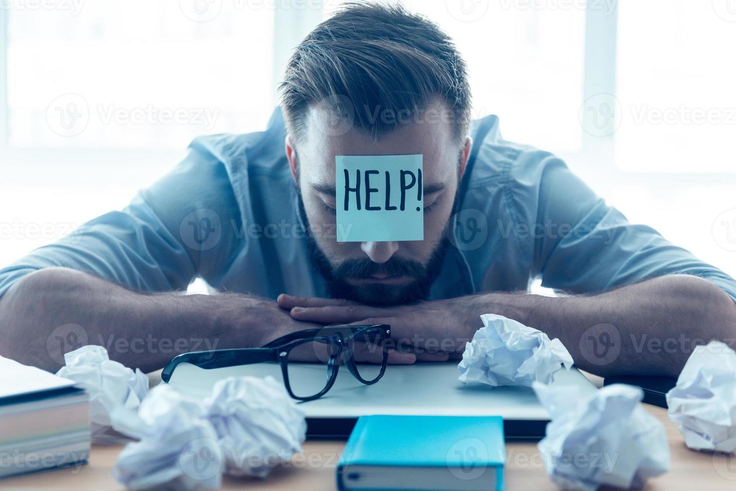 He needs help. Hopeless young beard man with adhesive note on his forehead leaning at the desk and keeping eyes closed while sitting at his working place in office photo