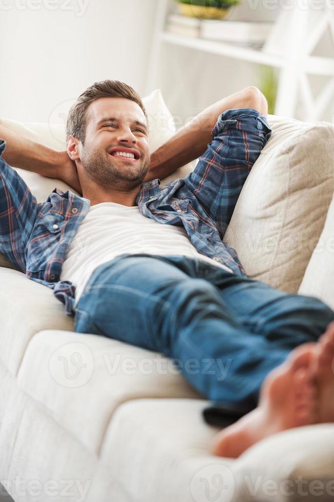 Enjoying lazy day at home. Happy young man holding hands behind head and smiling while lying on sofa photo
