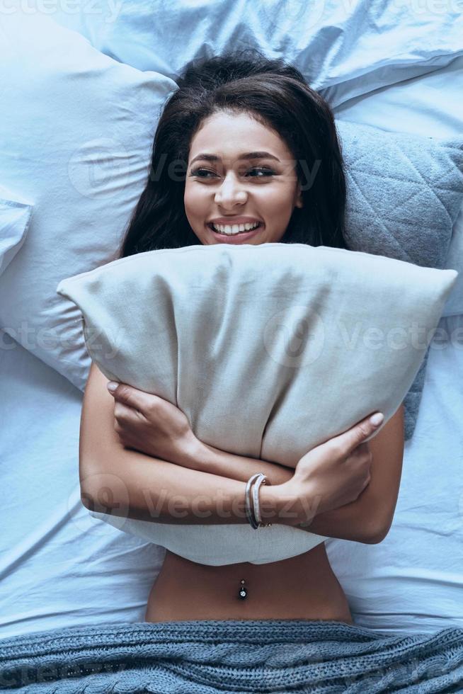 sentirse renovado y lleno de energía. vista superior de una atractiva joven abrazando la almohada y sonriendo mientras está acostada en la cama en casa foto