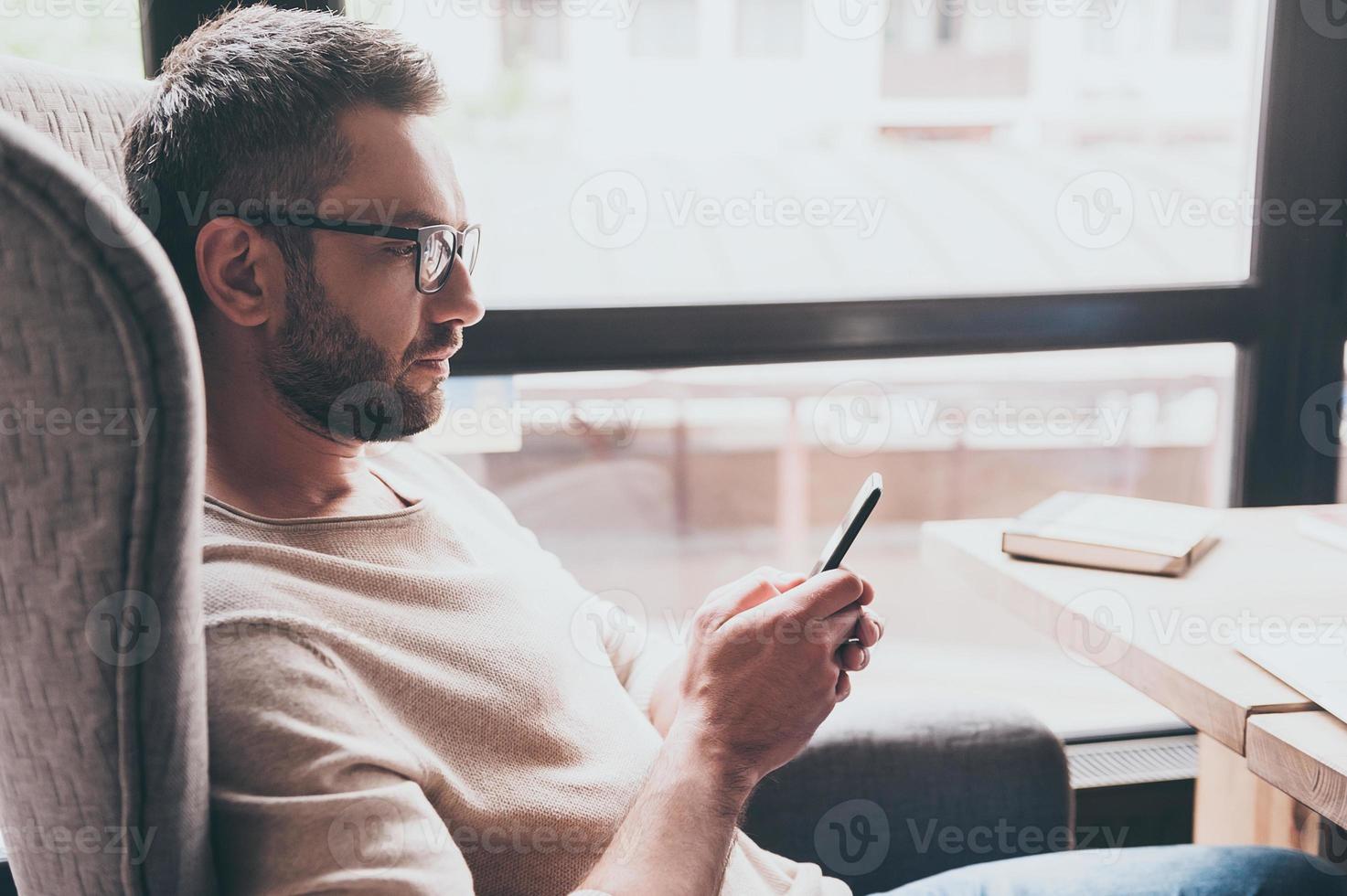 Business message. Side view of handsome man using his smartphone while sitting in chair in front of window photo
