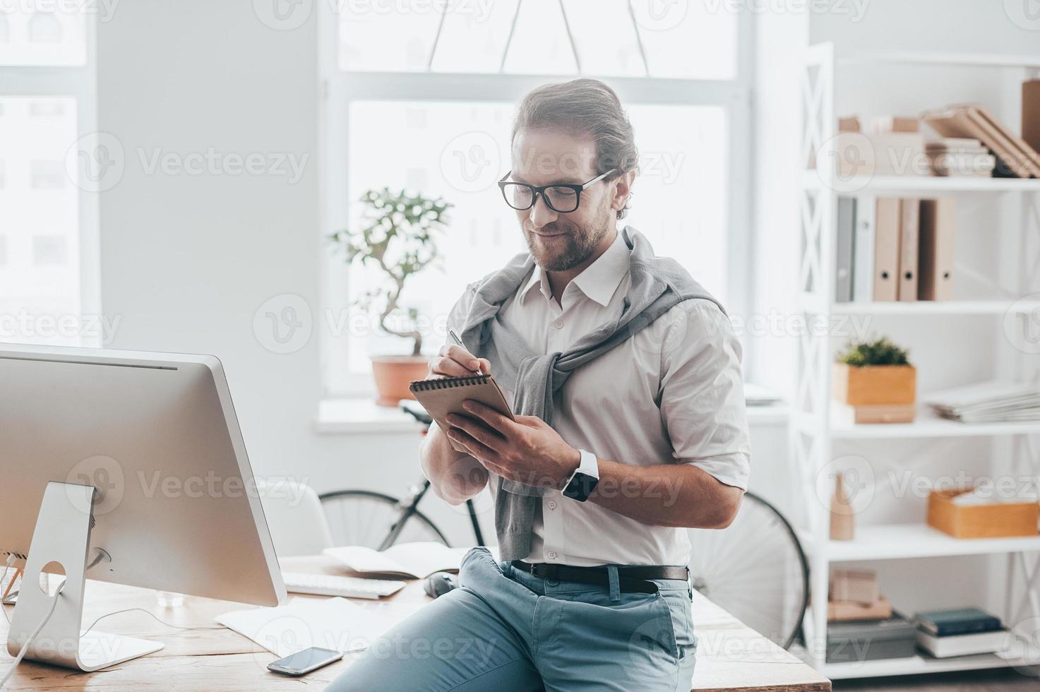 Thinking about new solutions. Handsome young man smiling and writing something while sitting on the desk at his working place in creative office photo