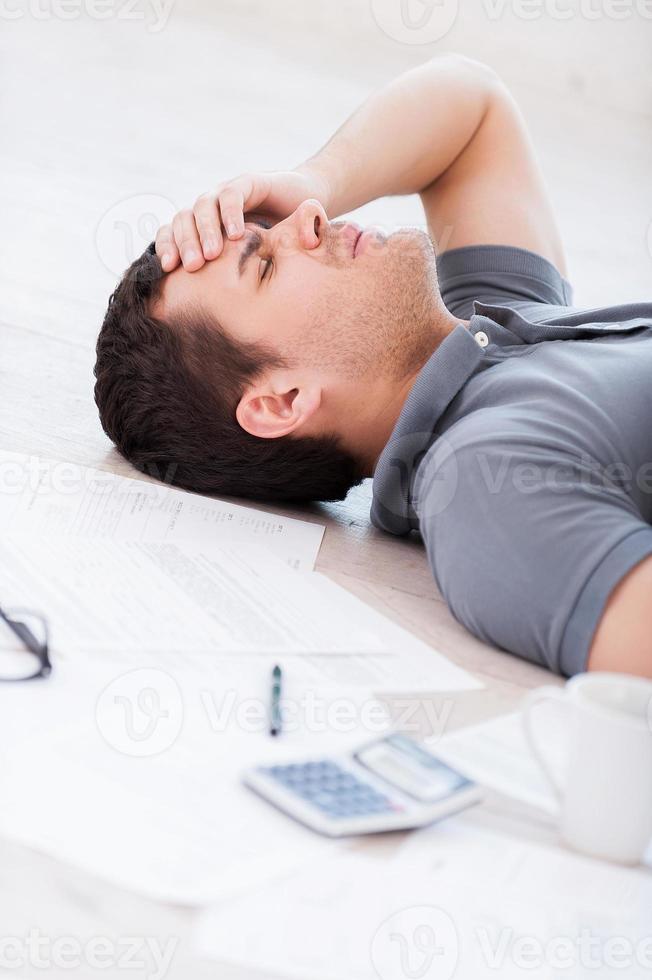 Overworked man. Top view of depressed young man holding hand on forehead while lying on the floor with documents and coffee cup on it photo