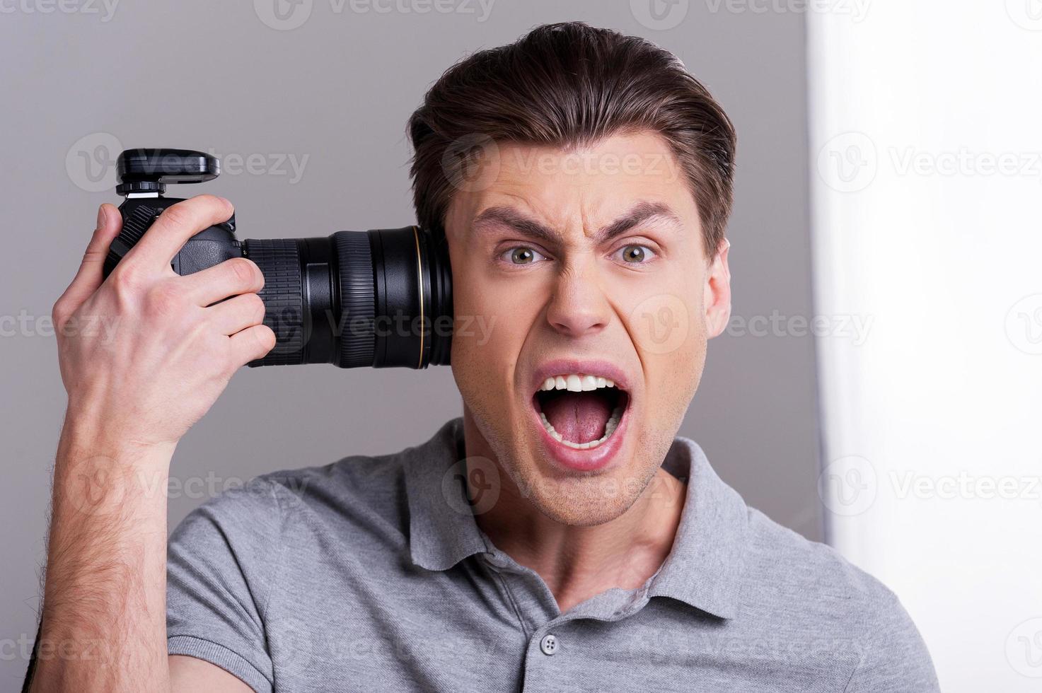 Me pegaré un tiro a mí mismo, un joven con un polo sosteniendo una cámara digital cerca de la cabeza y gritando mientras estoy de pie en el estudio con equipo de iluminación en segundo plano. foto