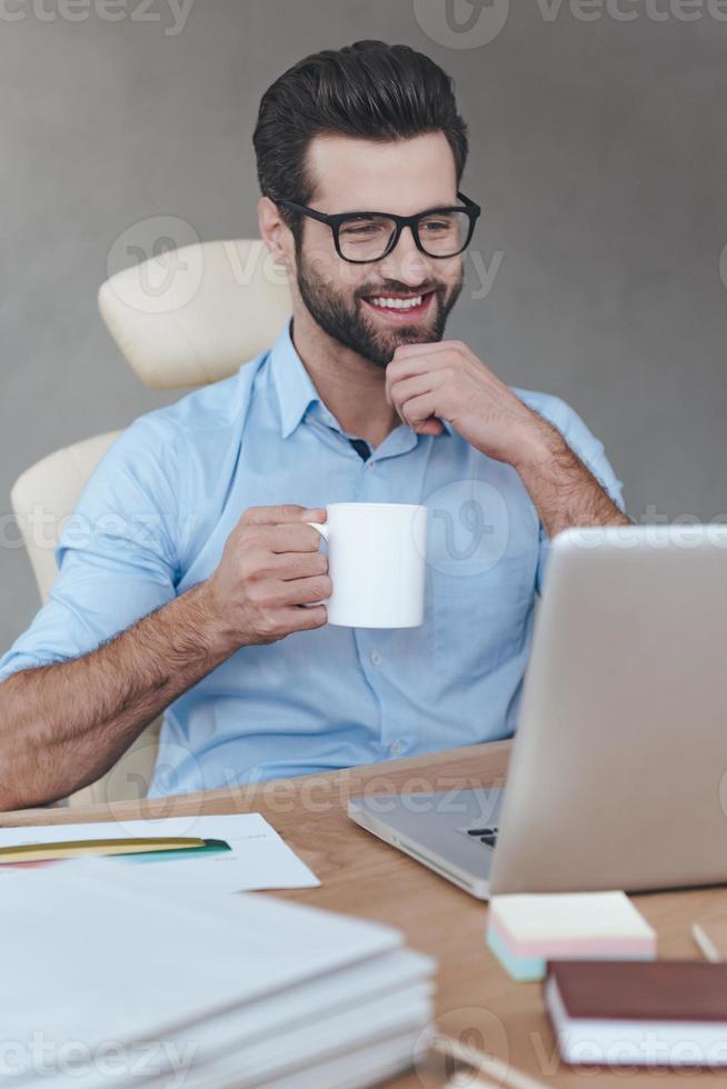 Coffee break. Cheerful young handsome man wearing glasses working on laptop with smile and holding coffee cup while sitting at his working place photo