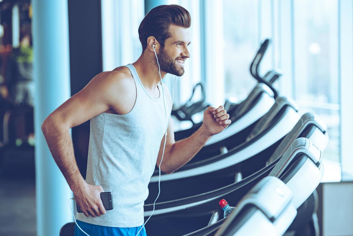 trotar con placer. vista lateral de un joven apuesto con auriculares mirando hacia otro lado con una sonrisa mientras corre en la cinta de correr en el gimnasio foto