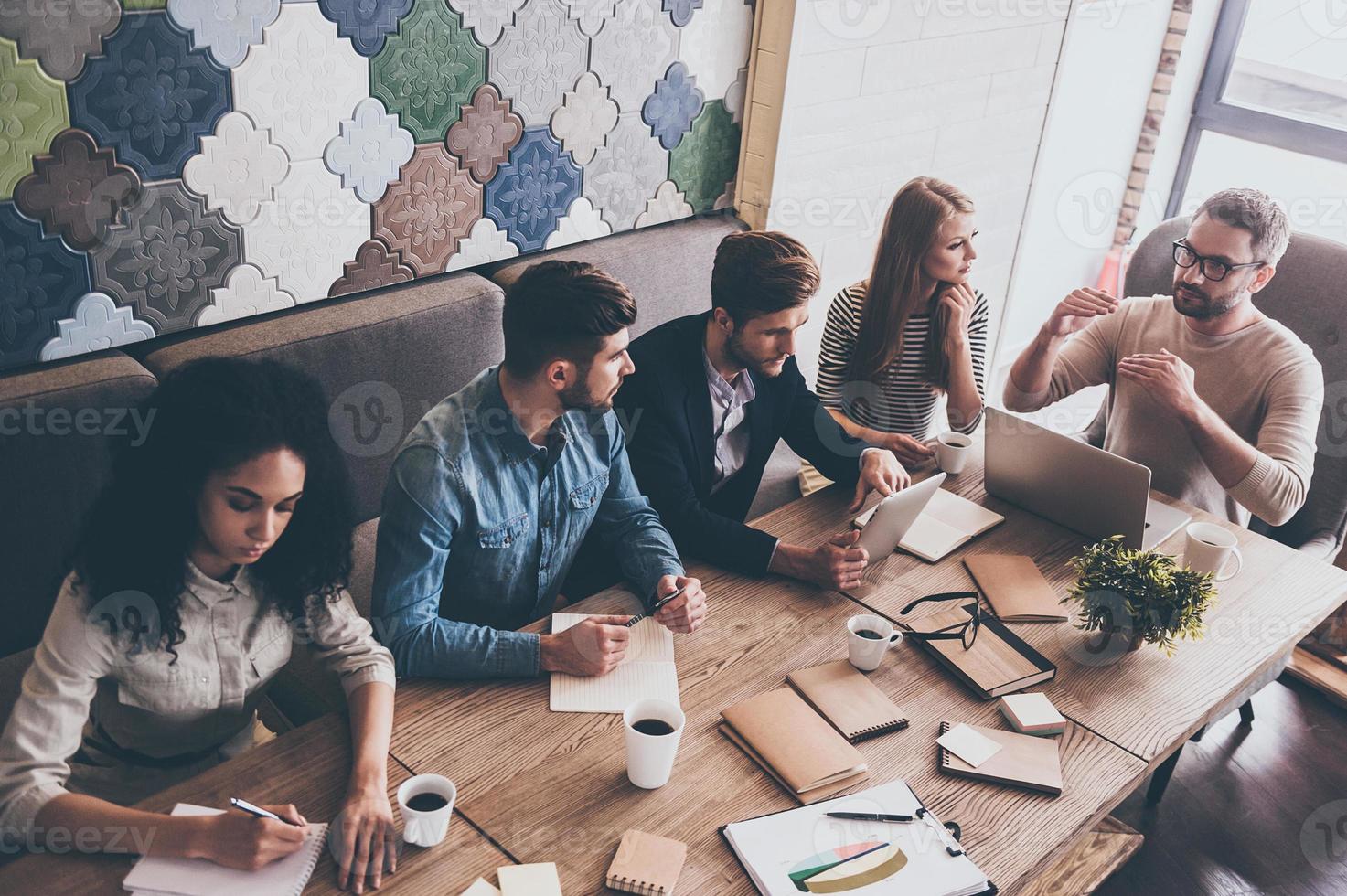 Combining their expertise. Top view of young handsome man in glasses gesturing and discussing something with his coworkers while sitting at the office table photo