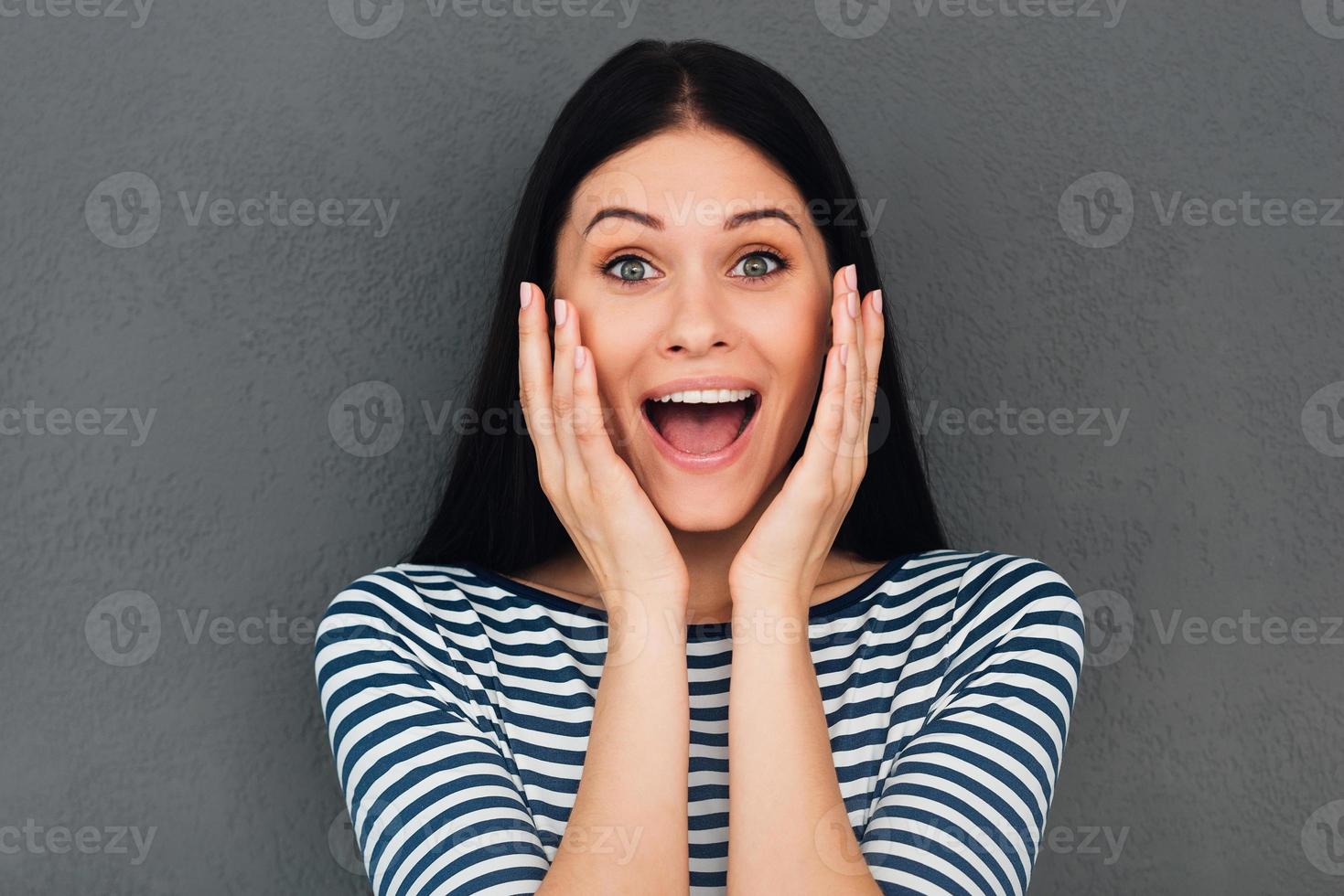 Wow Surprised young woman touching her face and smiling while standing against grey background photo