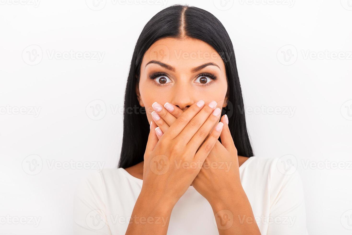 Shocking news. Surprised young woman covering mouth with hands and staring at camera while standing against white background photo
