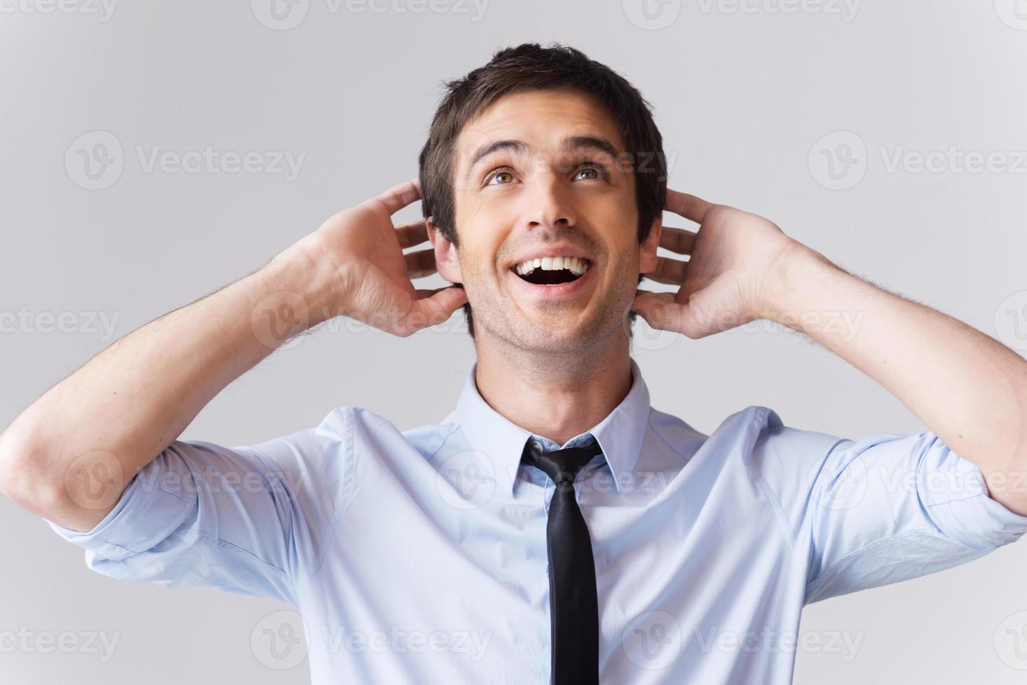 de ninguna manera emocionado joven con camisa y corbata tocando la cabeza con las manos y mirando hacia arriba con una sonrisa mientras está de pie contra el fondo gris foto