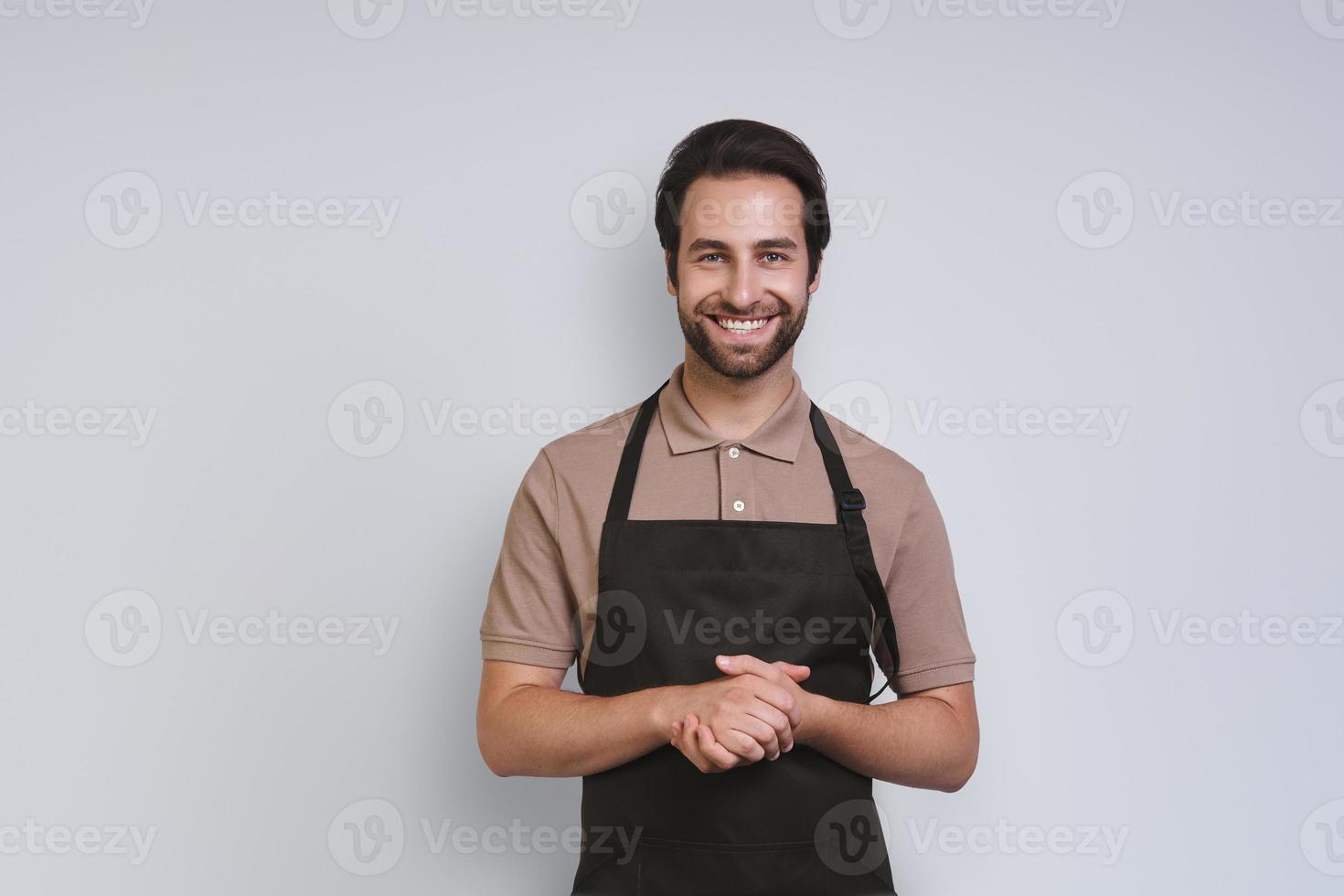 Handsome young man in apron looking at camera and keeping hands clasped while standing against gray background photo